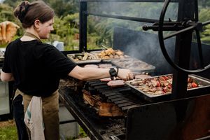 A chef grilling assorted meats on a large outdoor barbecue grill with smoke rising in a lush green setting.