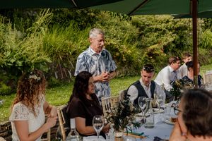 Man giving a speech at an outdoor wedding reception dinner table with guests listening and smiling.