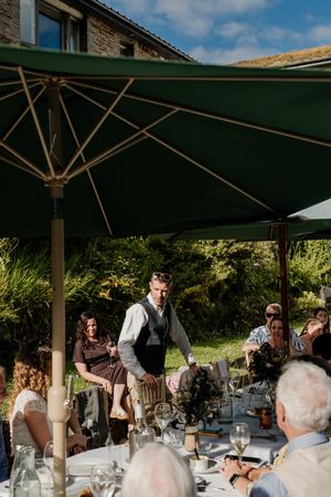 Groom stands at outdoor wedding reception table under green umbrella with guests seated around him.