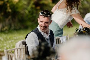 A smiling groom with sunglasses on his head sits at an outdoor wedding reception table.