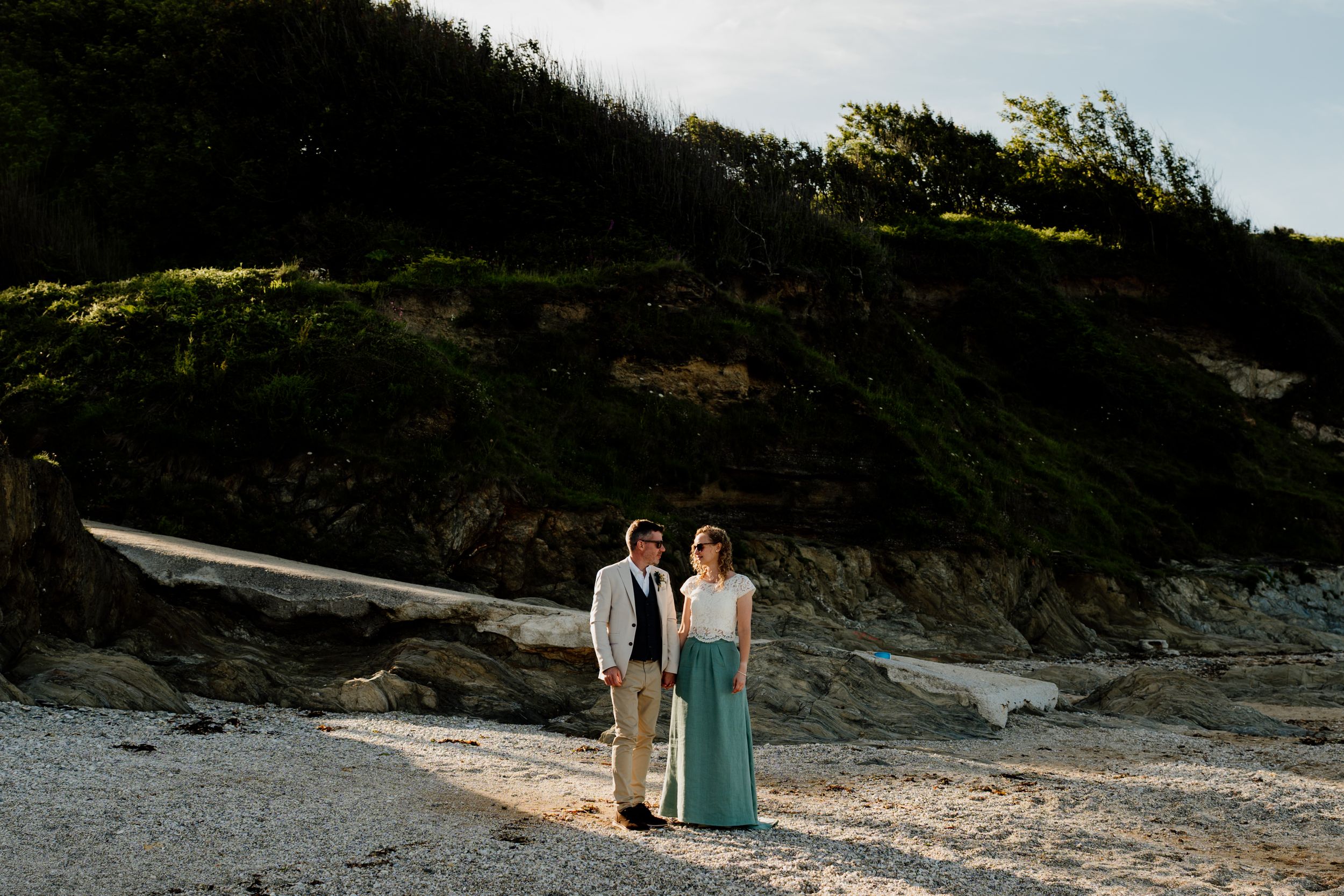 Couple standing on a rocky beach at sunset with green cliffs behind them during their wedding day.