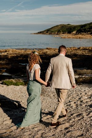 Bride and groom walk hand in hand along a rocky beach shoreline during their coastal wedding.