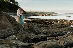 Couple embraces on rocky coastal beach at sunset during wedding photo session.