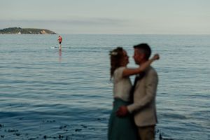 Blurred couple embracing on a rocky beach with a paddleboarder on calm blue sea in the background.