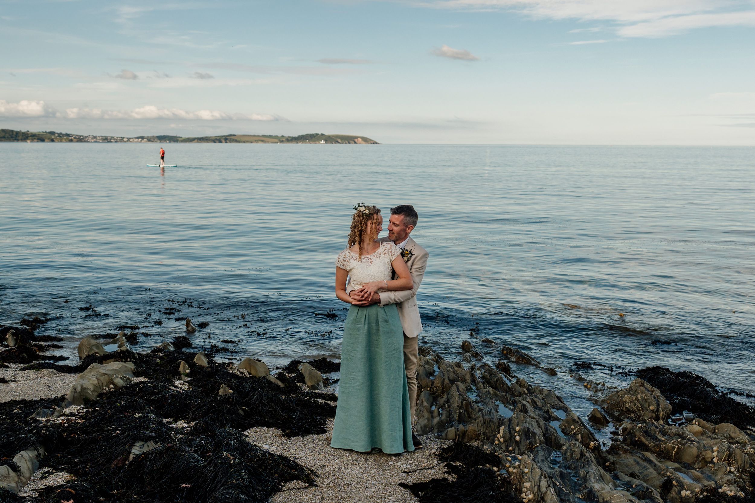 Couple embracing on rocky shoreline with calm ocean and coastal hills in background during wedding photo session.