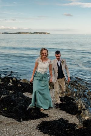 Bride in teal skirt and groom in tan suit walking over seaweed-covered rocks by the ocean.