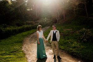 Bride and groom hold hands walking down sunlit forest path, bride wearing teal skirt.