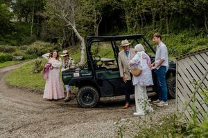Wedding guests gather around a green utility vehicle on a rural gravel path surrounded by lush greenery.
