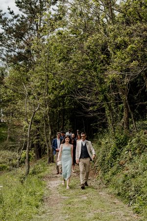 Wedding party walking hand-in-hand down a lush forest path on a sunny day.