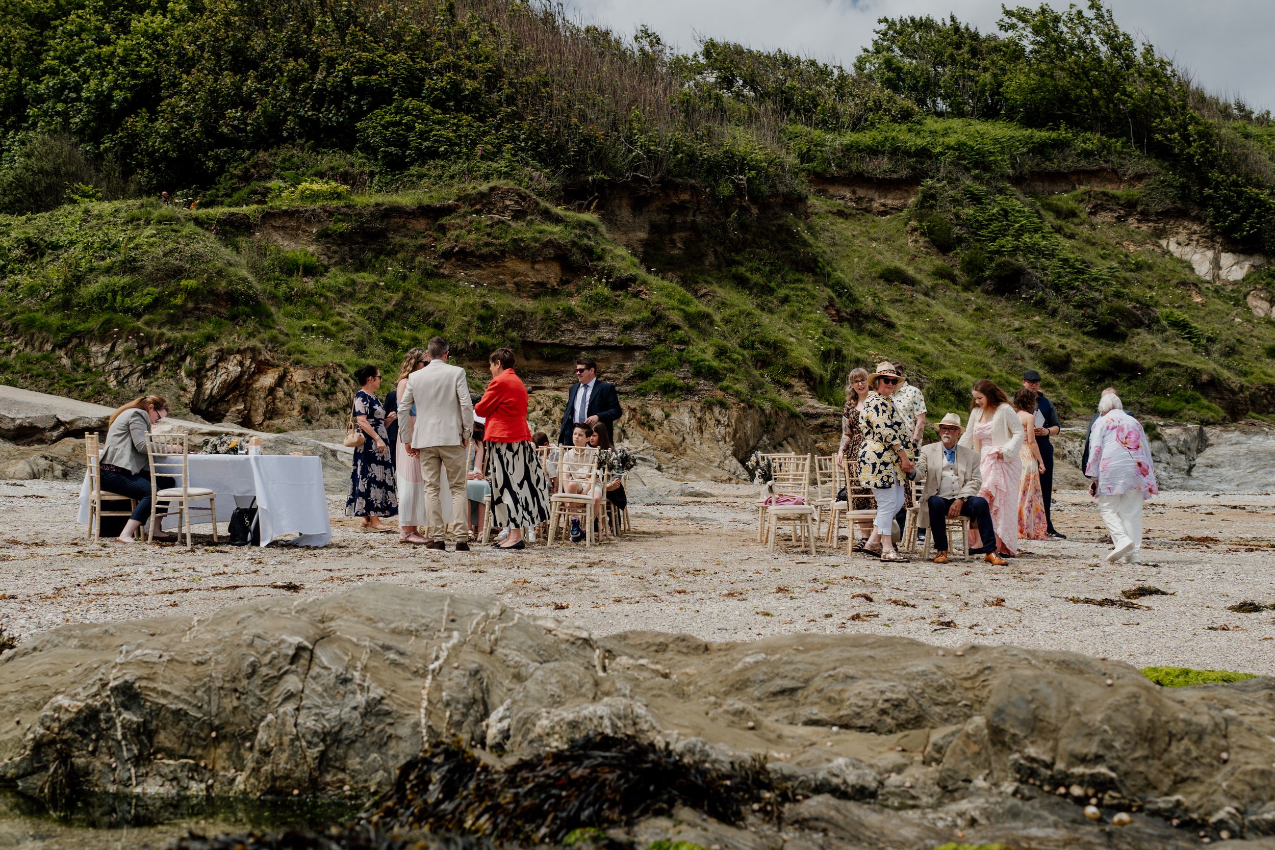 Small intimate beach wedding ceremony with guests gathered on sandy shore near green cliffs.