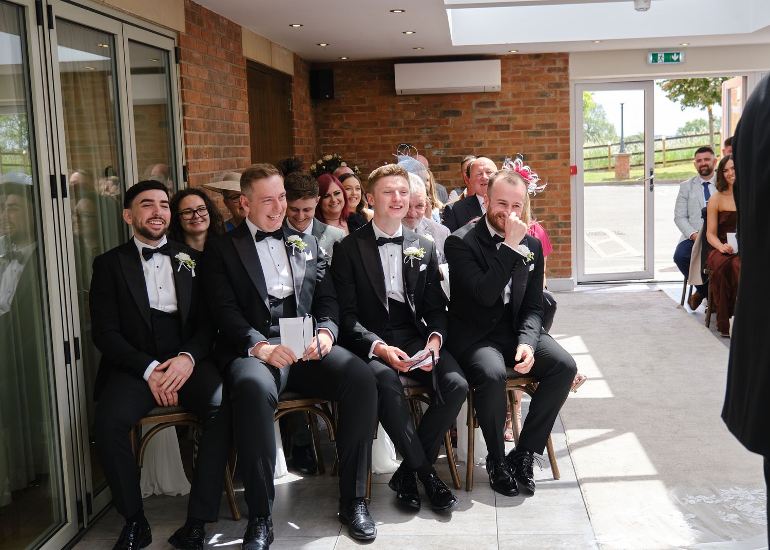 Wedding guests in black tuxedos seated and smiling during a ceremony in a modern venue with brick walls.