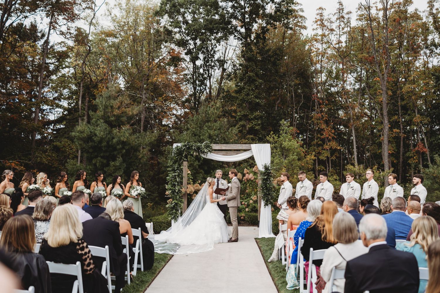 Outdoor wedding ceremony with bride and groom at floral arch, guests seated on white chairs, surrounded by lush green trees.