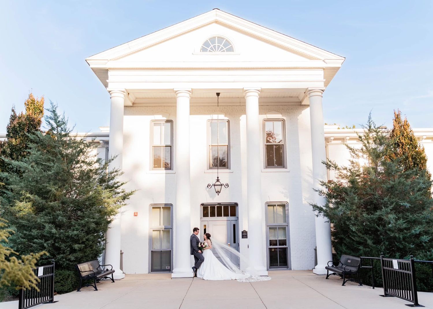 Bride and groom pose at entrance of elegant white colonial wedding venue with grand columns and lush trees.