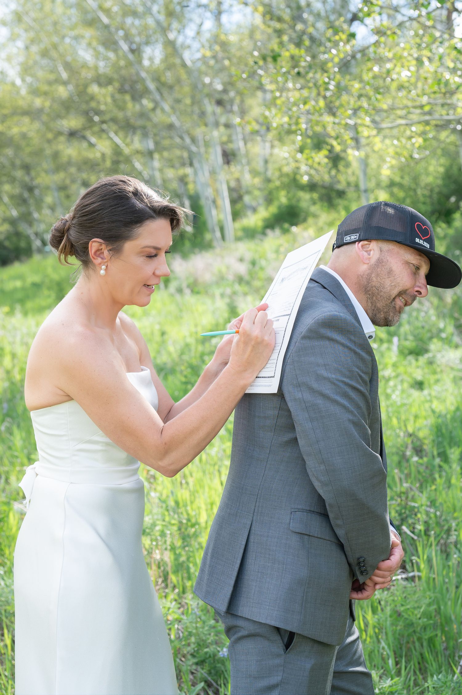 Bride in white dress signs marriage license on groom's back outdoors in lush green meadow during wedding ceremony at Vail elopement