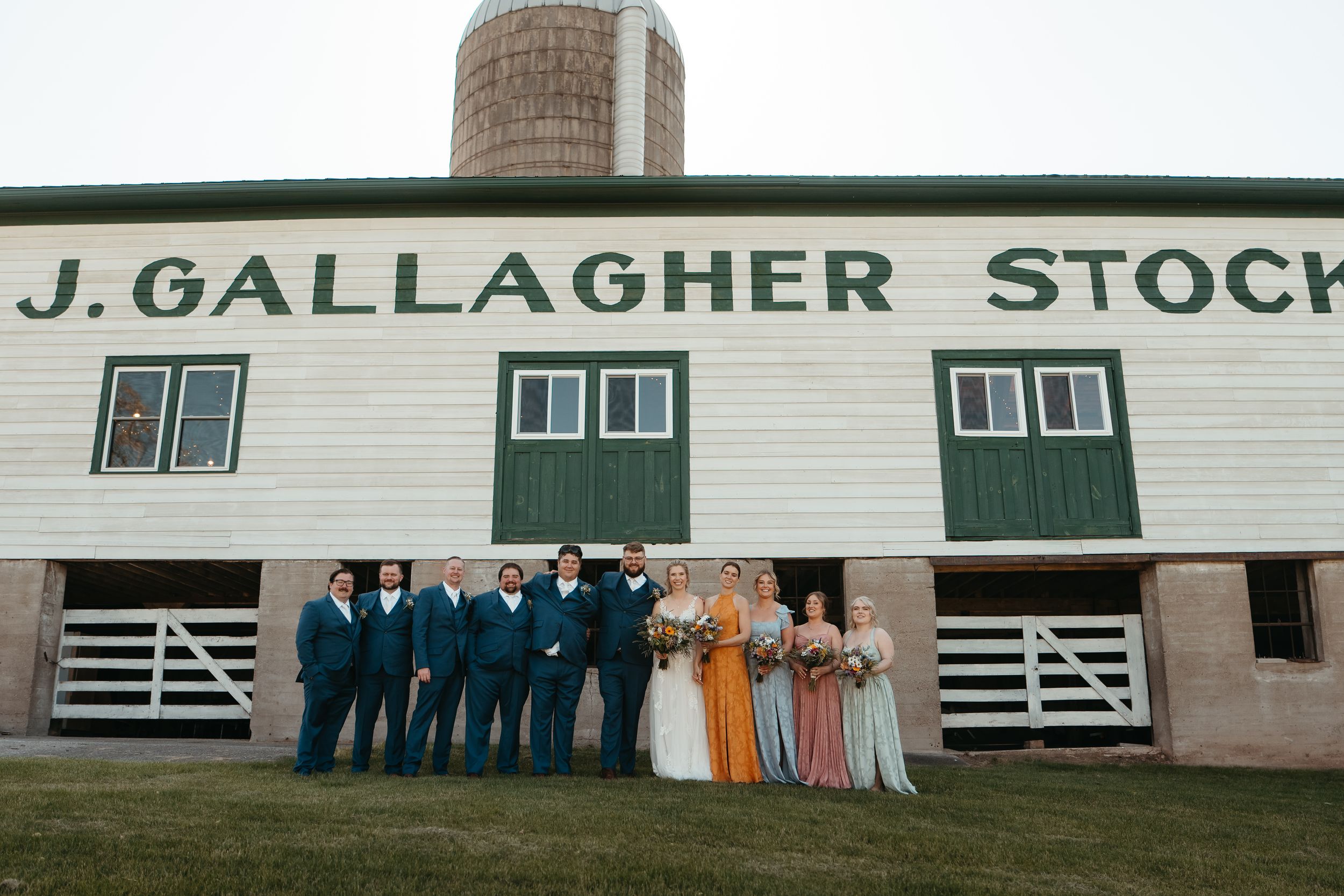 Wedding party poses outside J. Gallagher Stock barn, groomsmen in teal suits, bridesmaids in floral gowns, bride in white.