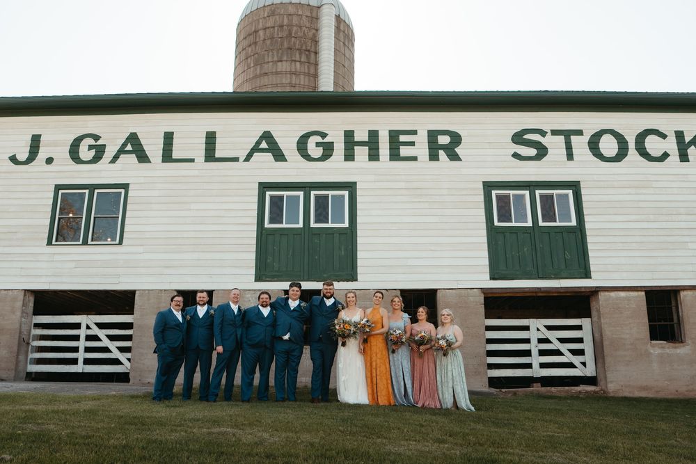 Wedding party poses outside J. Gallagher Stock barn, groomsmen in teal suits, bridesmaids in floral gowns, bride in white.