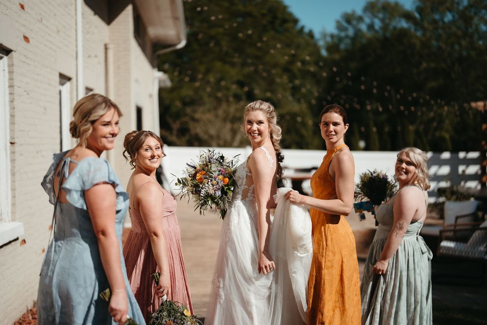 Bride in white gown poses with bridesmaids in colorful mismatched dresses holding bouquets outdoors.