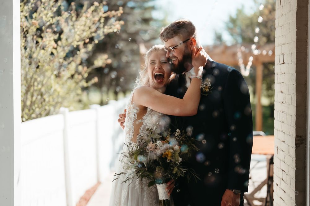 Happy bride and groom embracing outdoors with confetti, bride holding wildflower bouquet in elegant wedding attire.