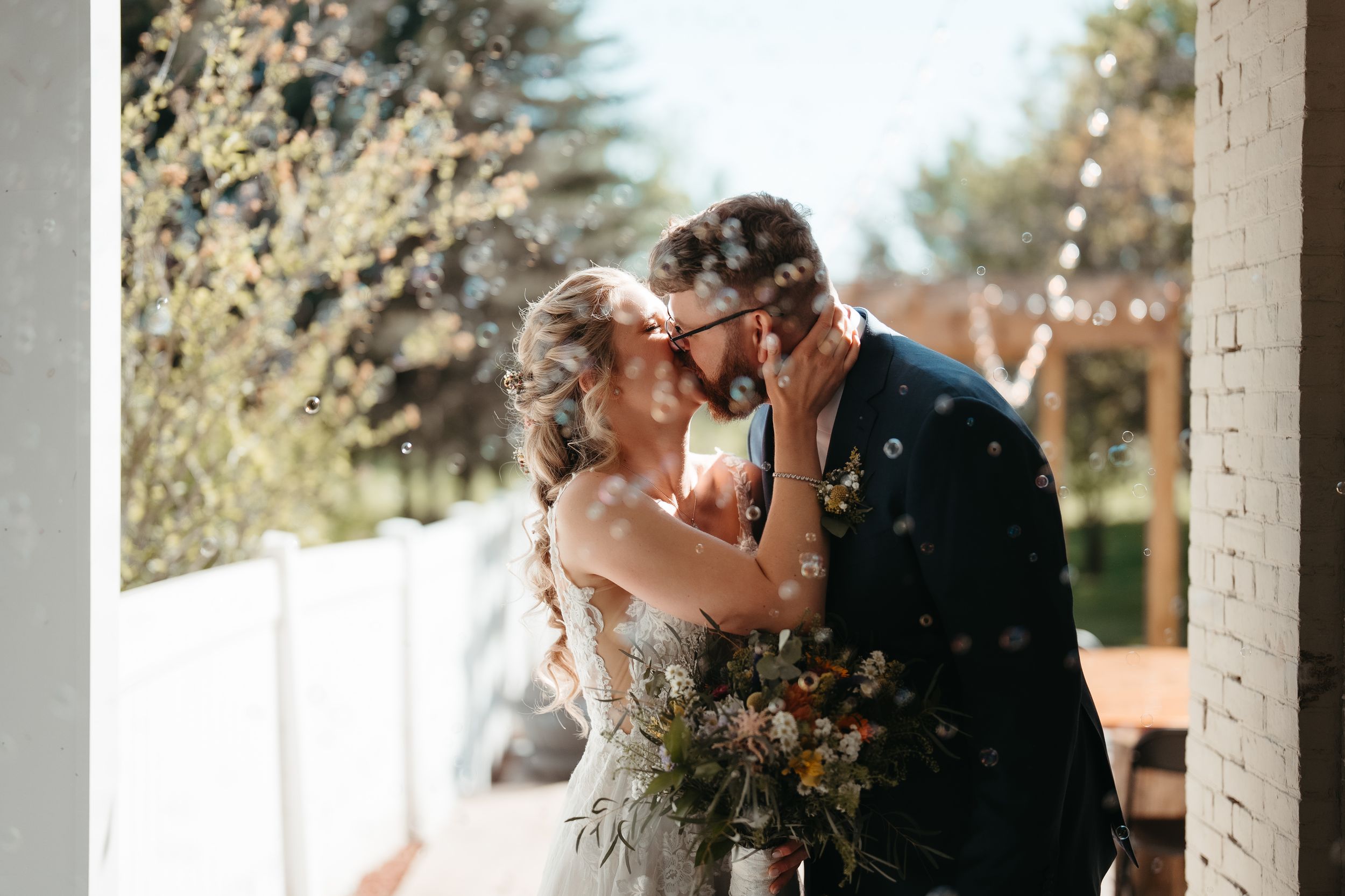Bride and groom sharing a kiss outdoors with confetti falling, bride holding a floral bouquet on their wedding day.