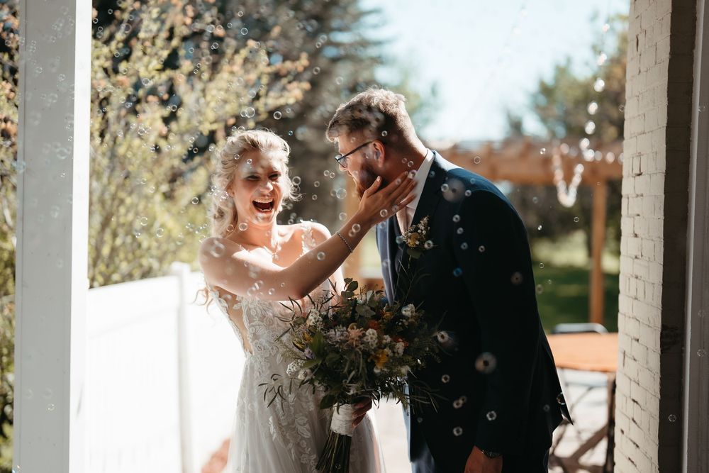 Joyful bride and groom laughing together outdoors, surrounded by confetti, bride holding a rustic bouquet on their wedding day.