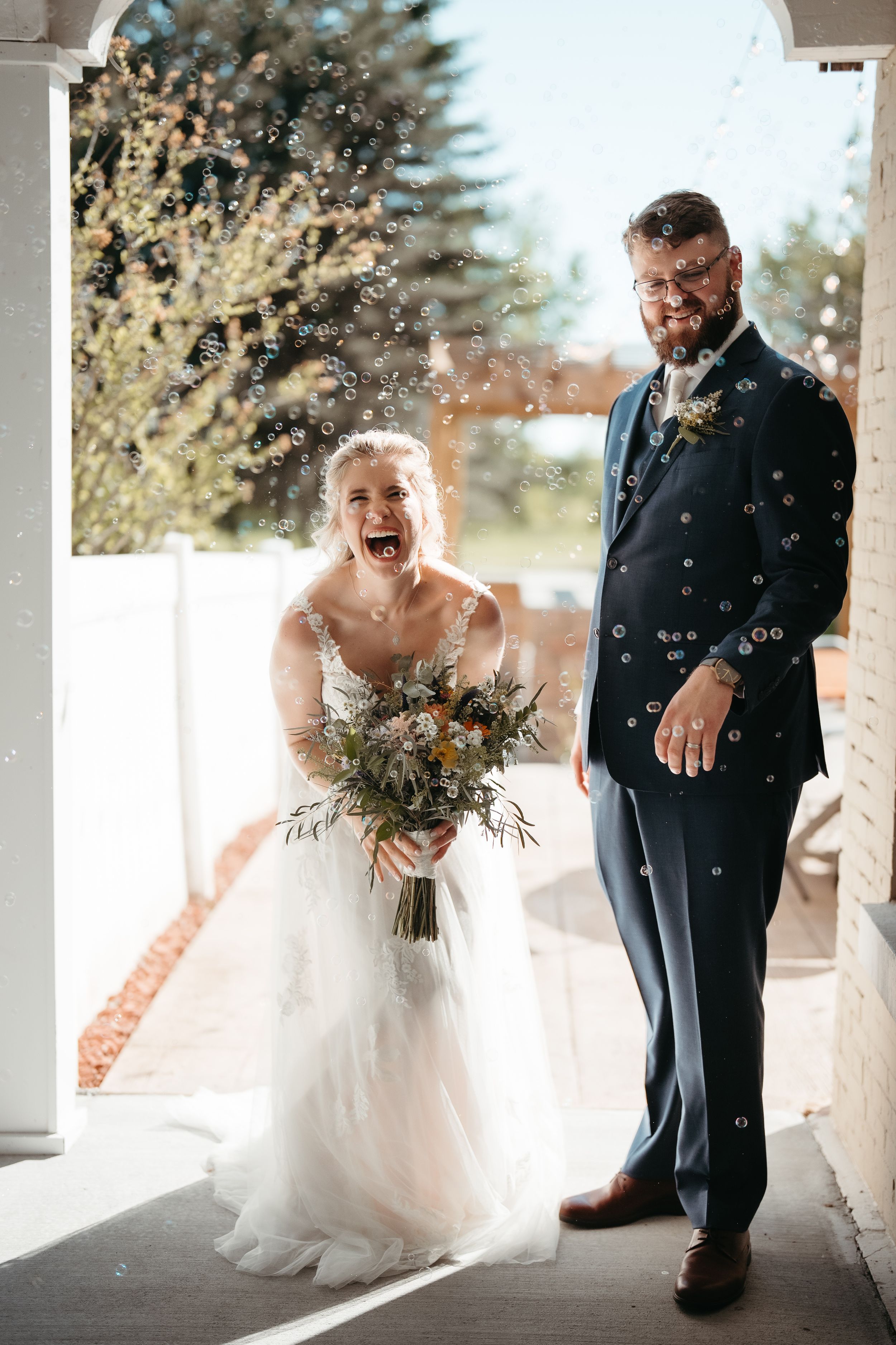 Joyful bride and groom laughing as bubbles float around them during outdoor wedding celebration, bride holding wildflower bouquet.