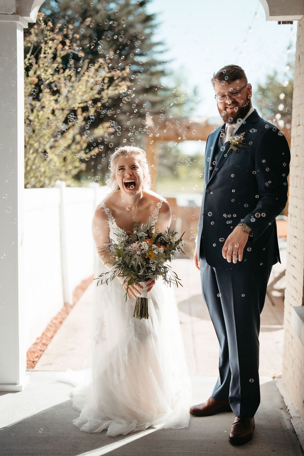 Joyful bride and groom laughing as bubbles float around them during outdoor wedding celebration, bride holding wildflower bouquet.