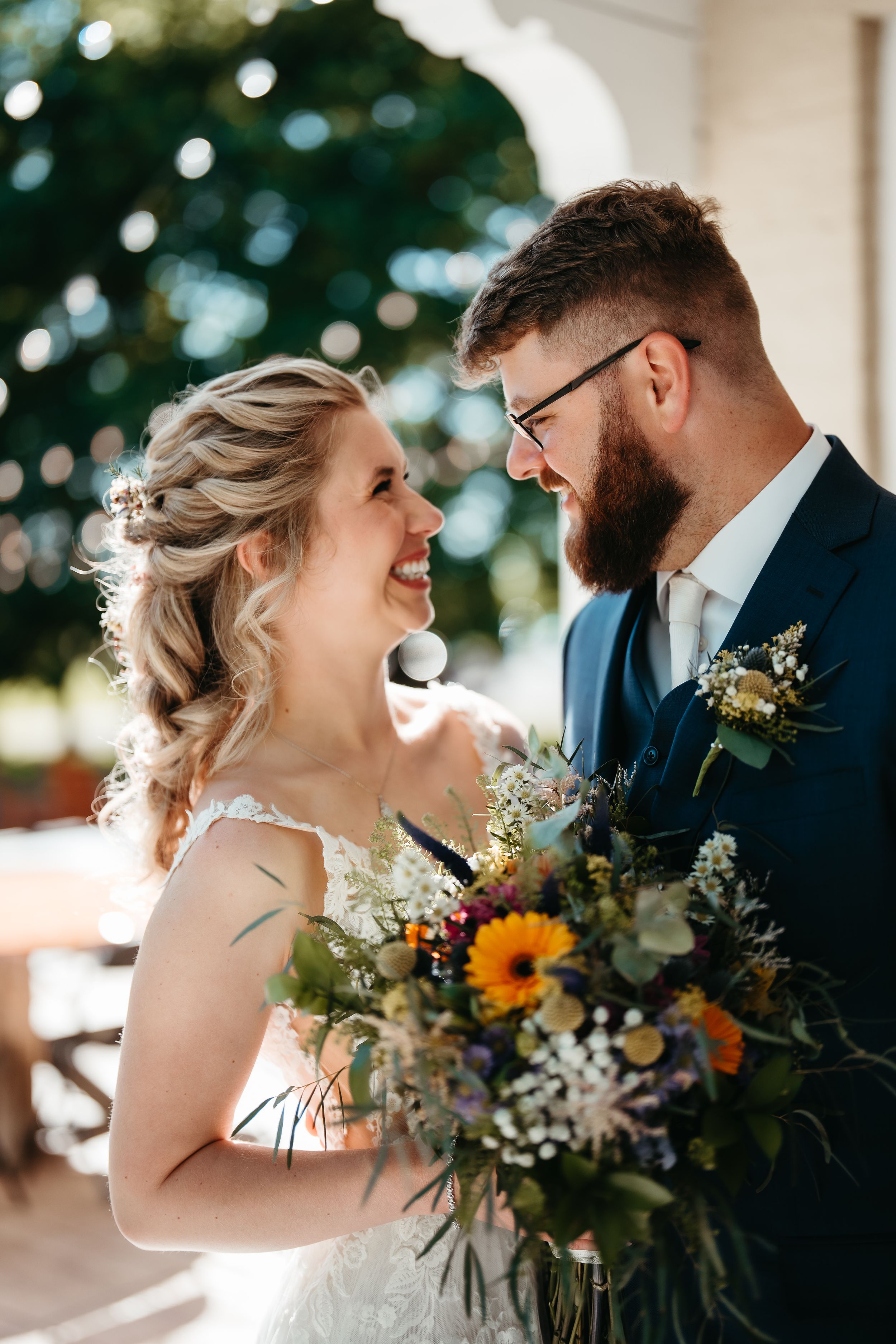 Smiling bride holding colorful wildflower bouquet gazes at bearded groom in navy suit at outdoor wedding.