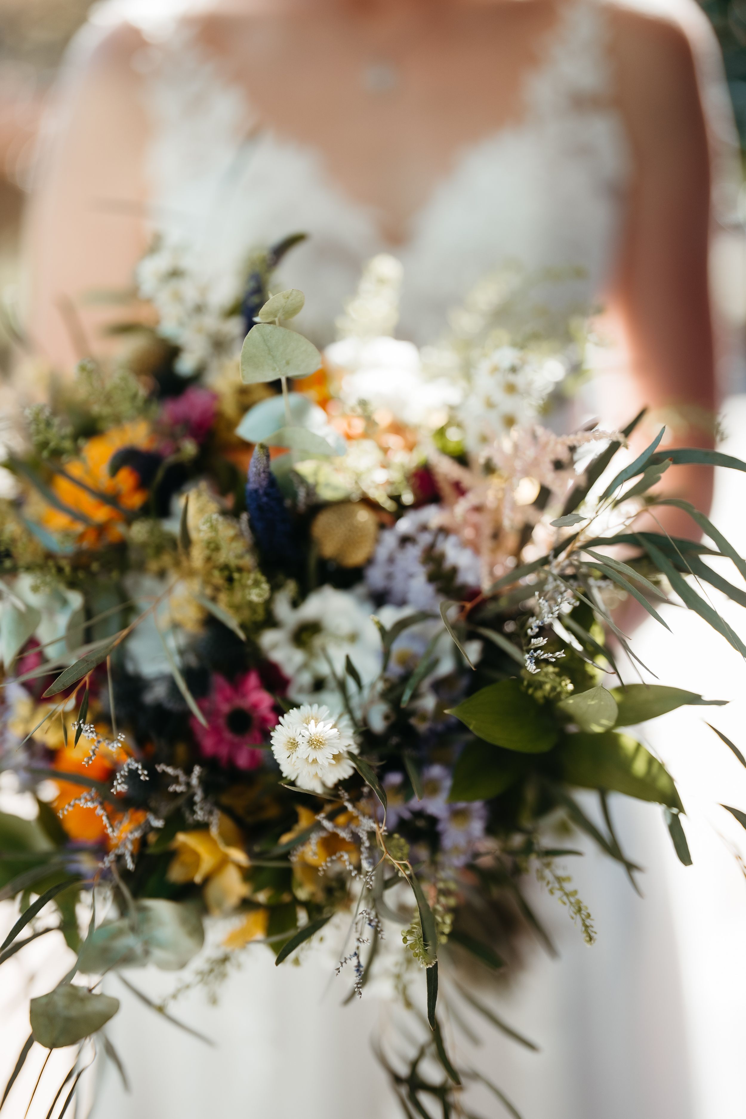 Bride holding a lush wildflower wedding bouquet with sunflowers, daisies, and greenery in vibrant fall colors.