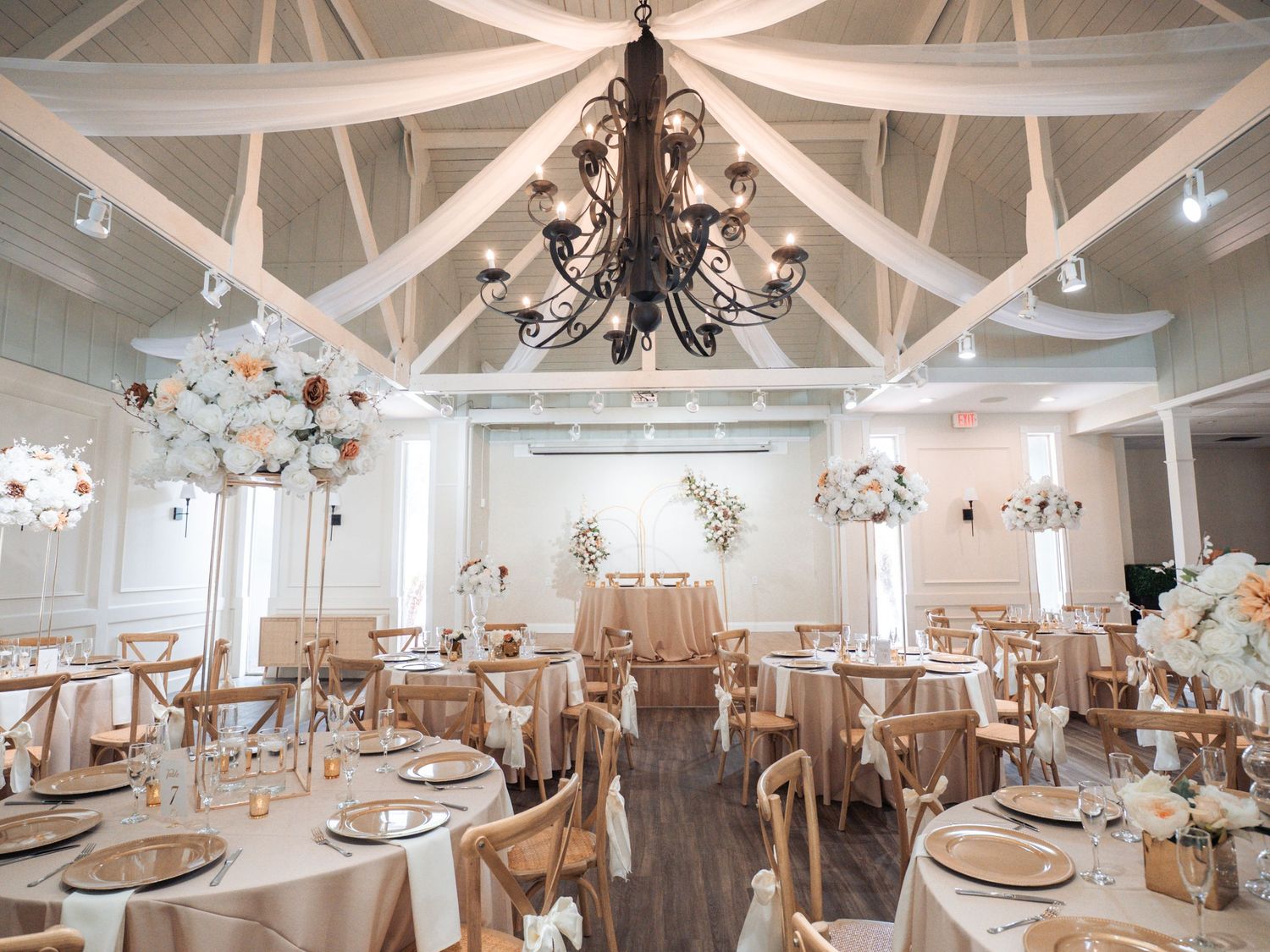 Elegant wedding reception hall with chandelier, draped ceilings, round tables with champagne linens, and floral centerpieces.