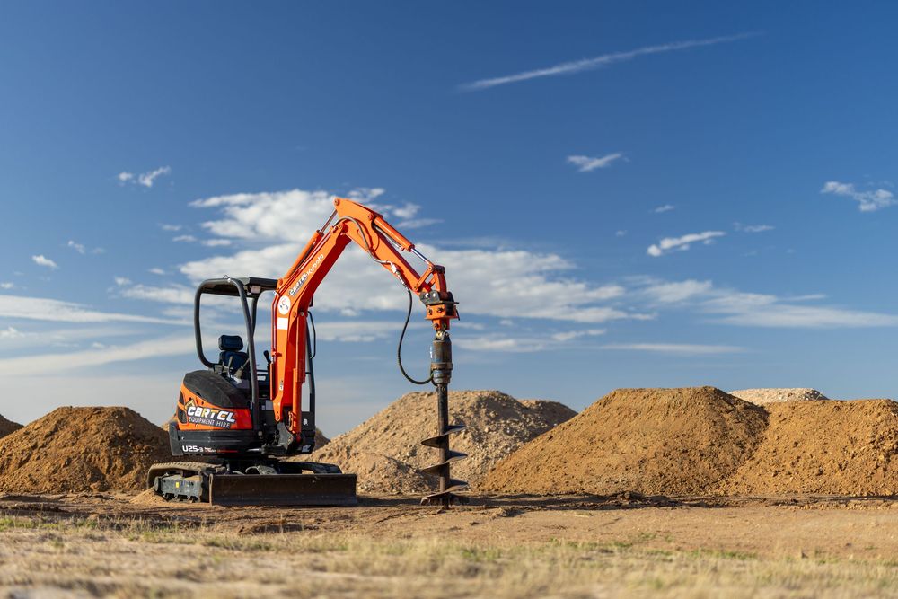 Orange mini excavator with auger attachment working near dirt mounds under a blue sky.