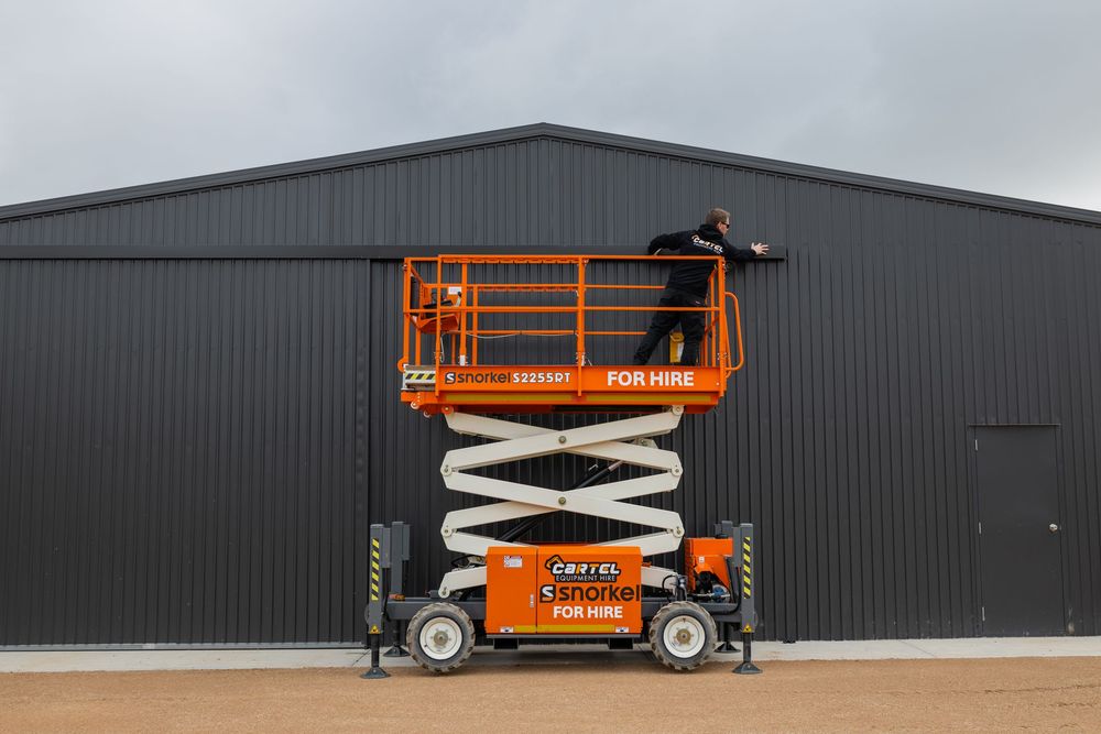 Orange scissor lift for hire positioned against a dark grey industrial warehouse building outdoors.