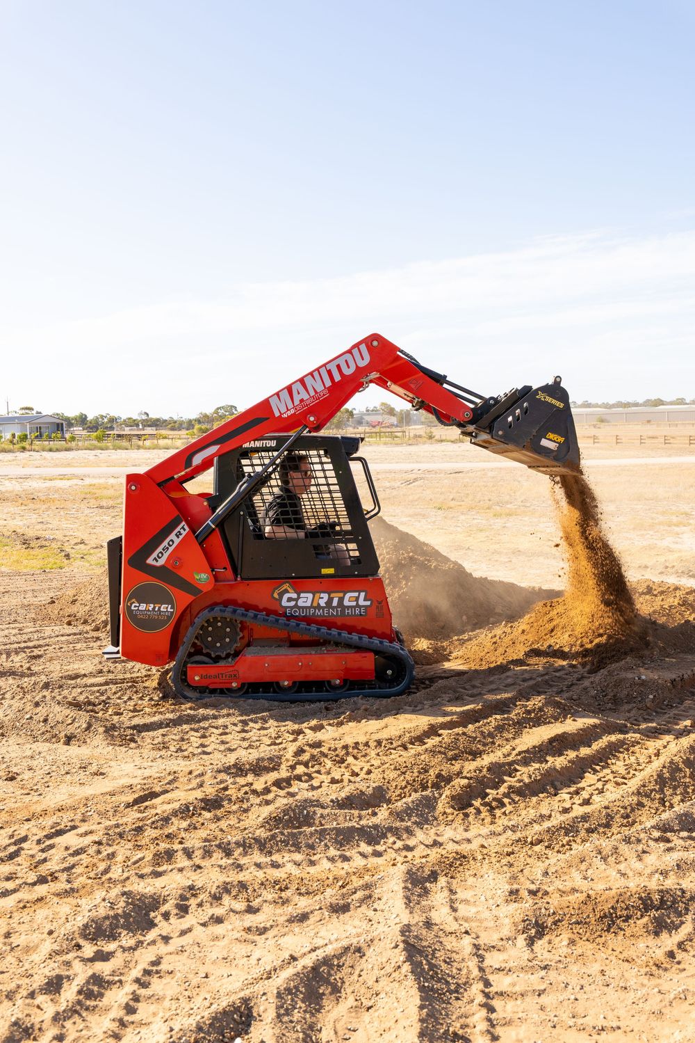 Red Manitou skid steer loader dumping dirt on a construction site under a clear blue sky.