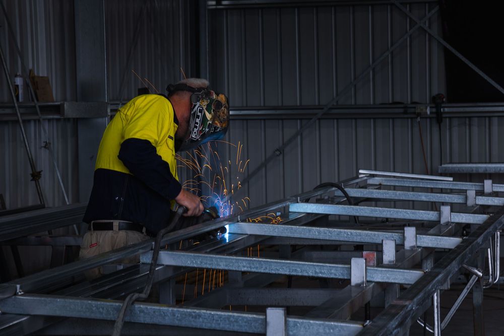 Worker in hi-vis welding a steel frame structure inside an industrial shed with sparks flying.