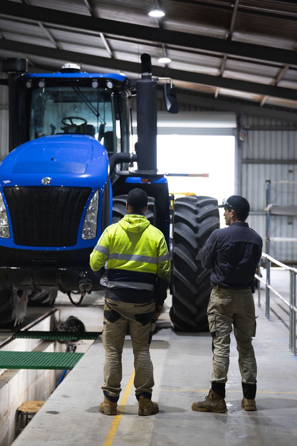Two workers inspecting a large blue New Holland tractor in a shed.