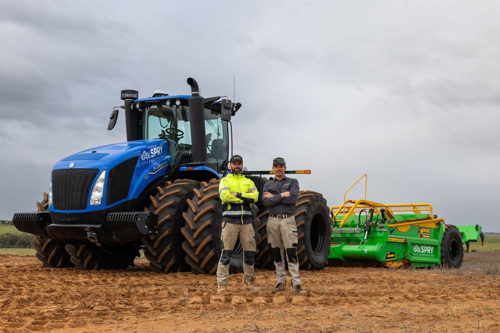 Two workers in hi-vis standing before a large blue New Holland tractor with green equipment.