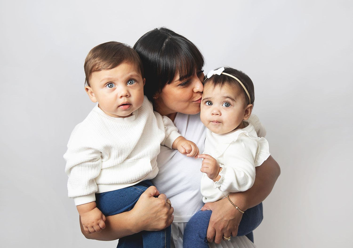 A mother kisses one of her twin babies while holding both infants dressed in whit during a family photoshoot in Bexley