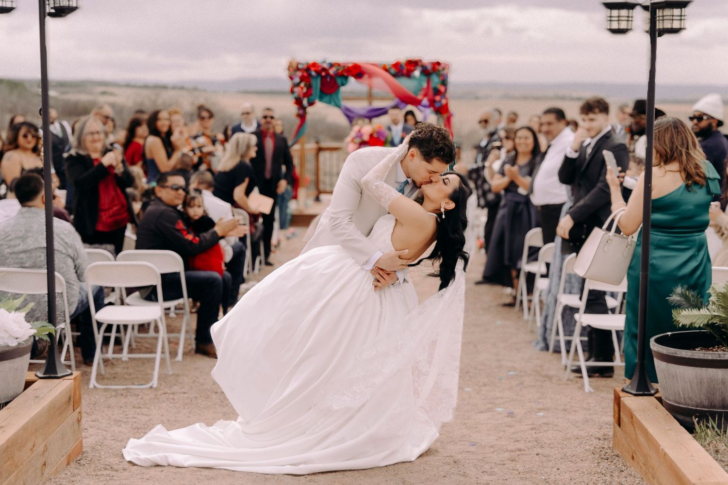 Groom dips bride for a kiss at outdoor wedding ceremony with guests cheering in background.