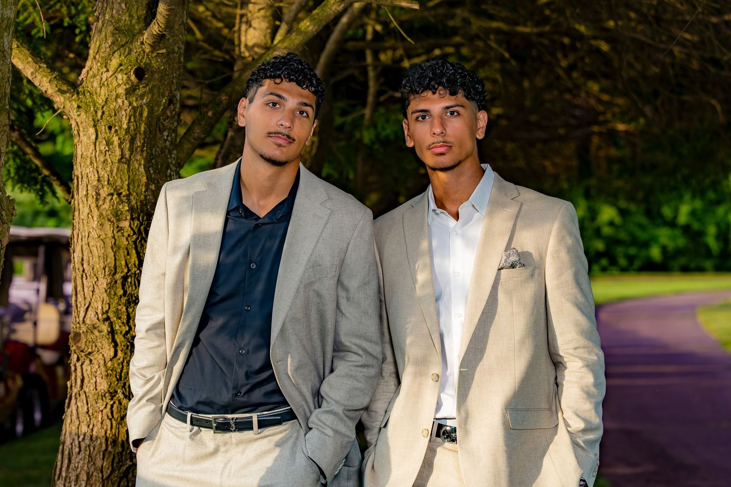 Two young men in light gray suits pose outdoors near a tree on a lush green path.