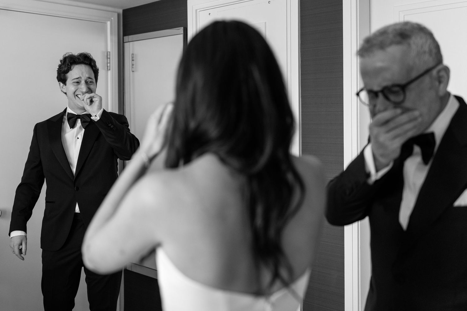 Emotional groom and father react with tears seeing bride in wedding dress, captured in black and white photography.