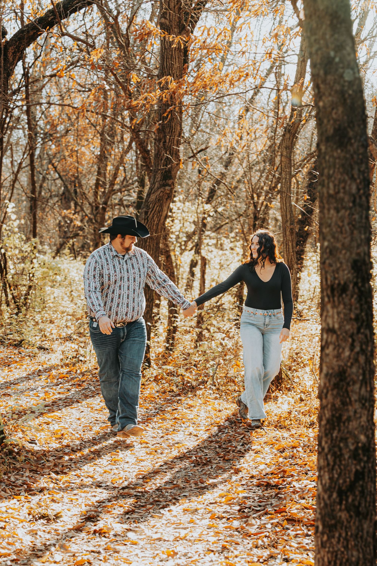 Couple holding hands walking on a leaf-covered autumn forest path, man in cowboy hat and woman in black top.