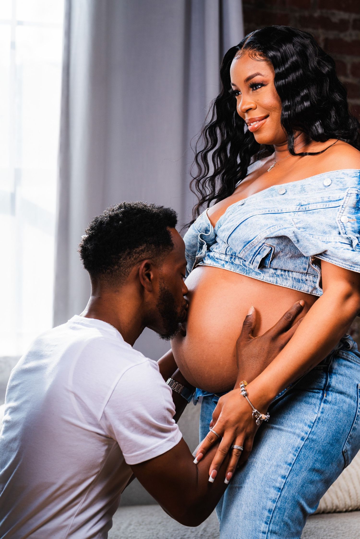 A man kissing a pregnant woman's belly during a maternity photoshoot, both dressed in denim outfits.