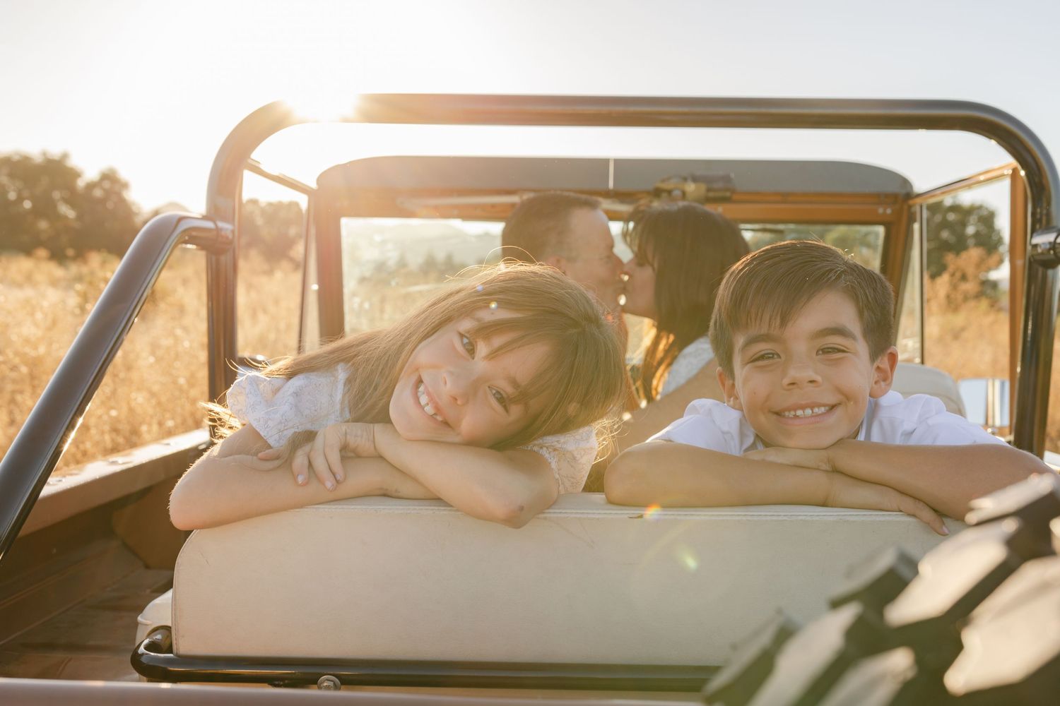 Two happy children lean over the back seat of an open-top jeep in a sunny golden field.