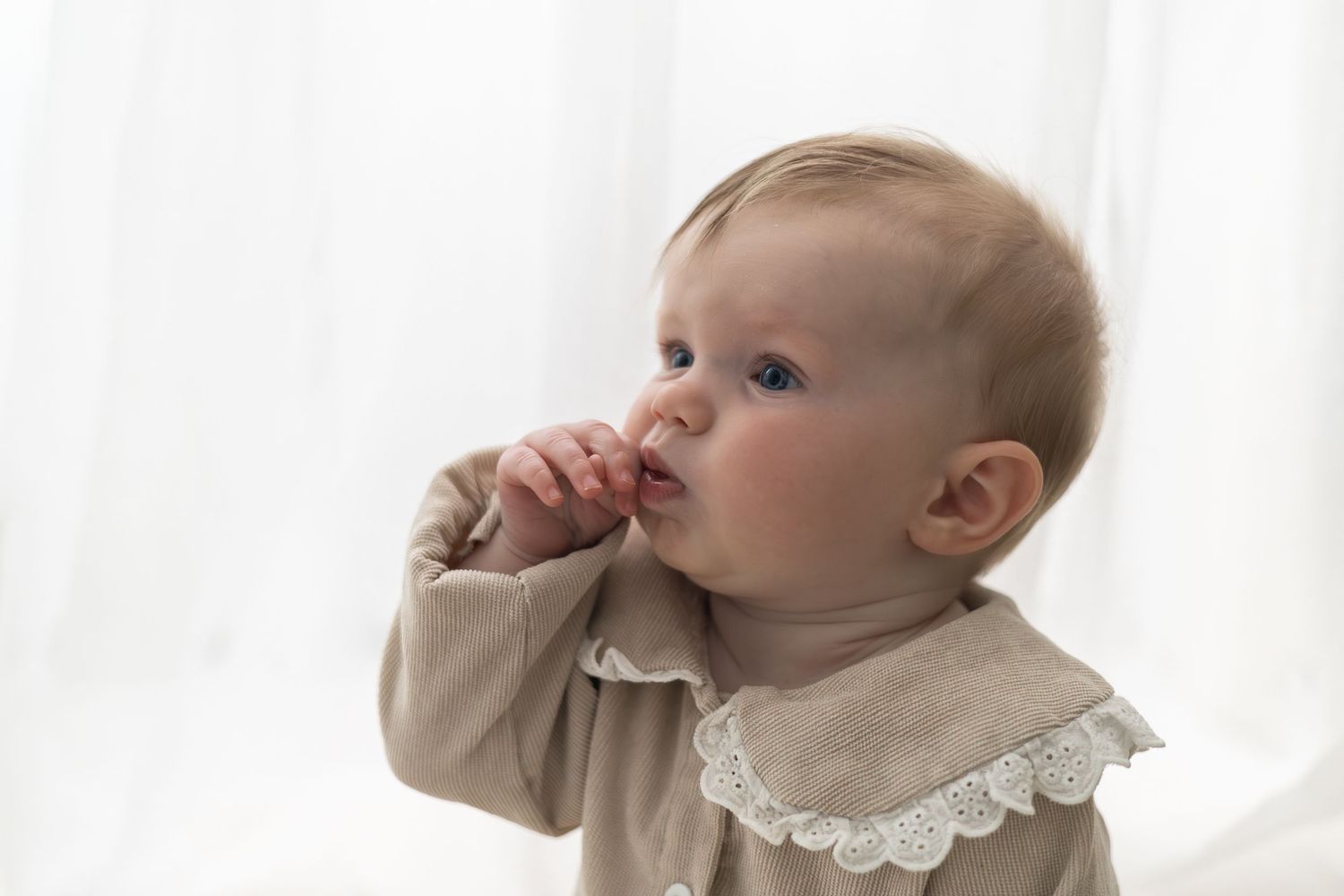 Adorable baby in beige onesie with fingers near mouth, soft white background, Birthday photographer Nottingham