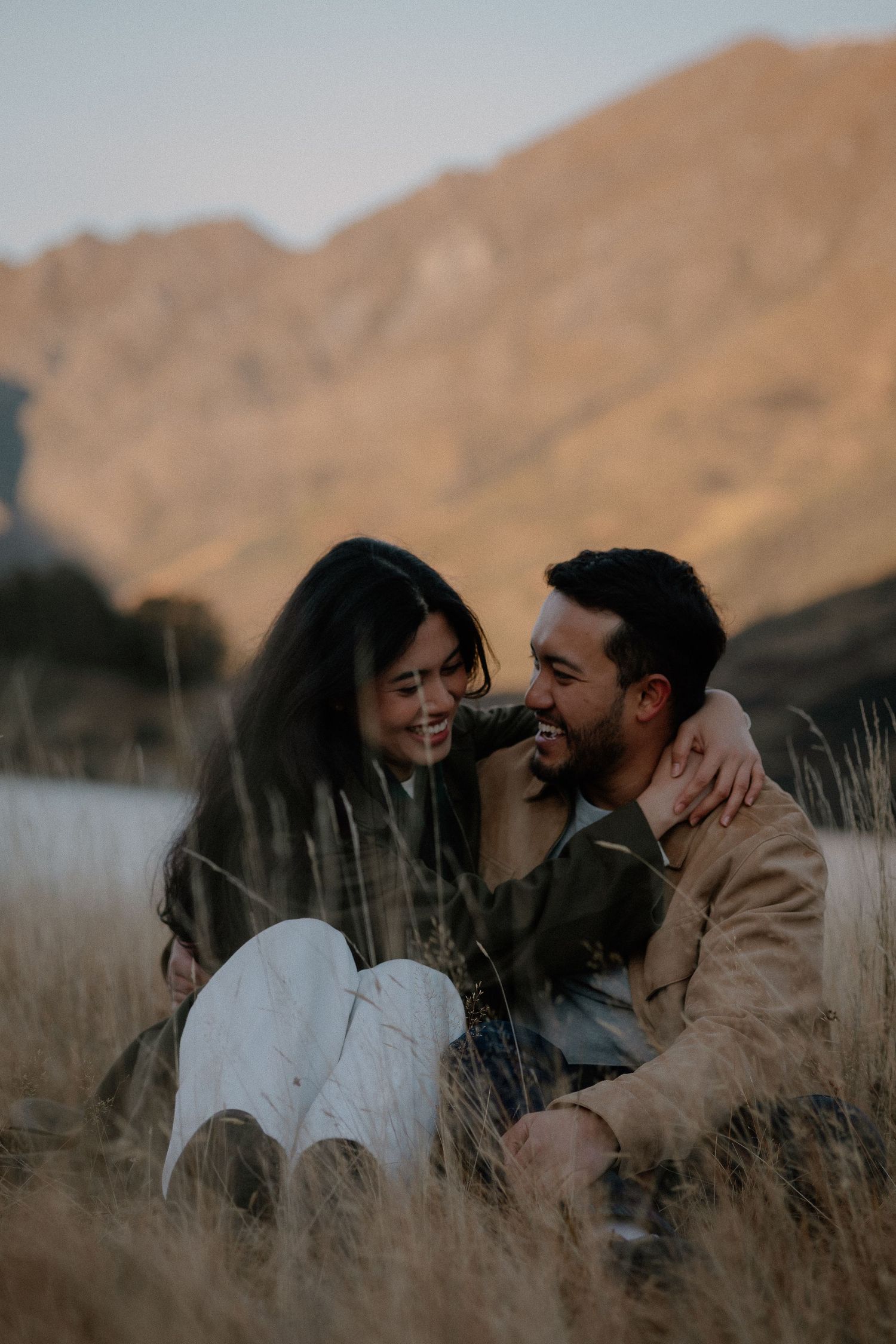 A happy couple sits together in golden grass, laughing and embracing with a sunlit mountain backdrop.
