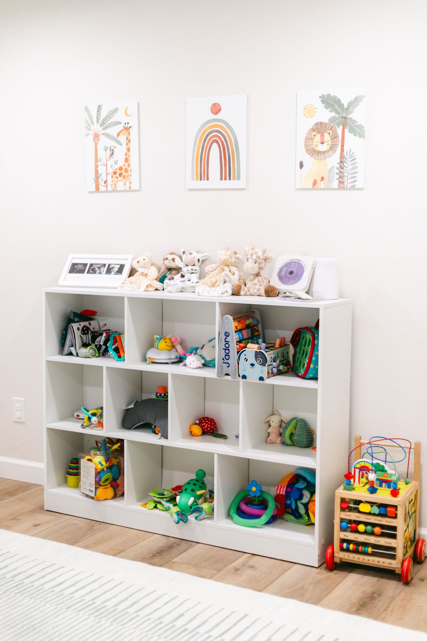 White cubby shelf filled with colorful toys and stuffed animals in a bright nursery with safari-themed wall art above.