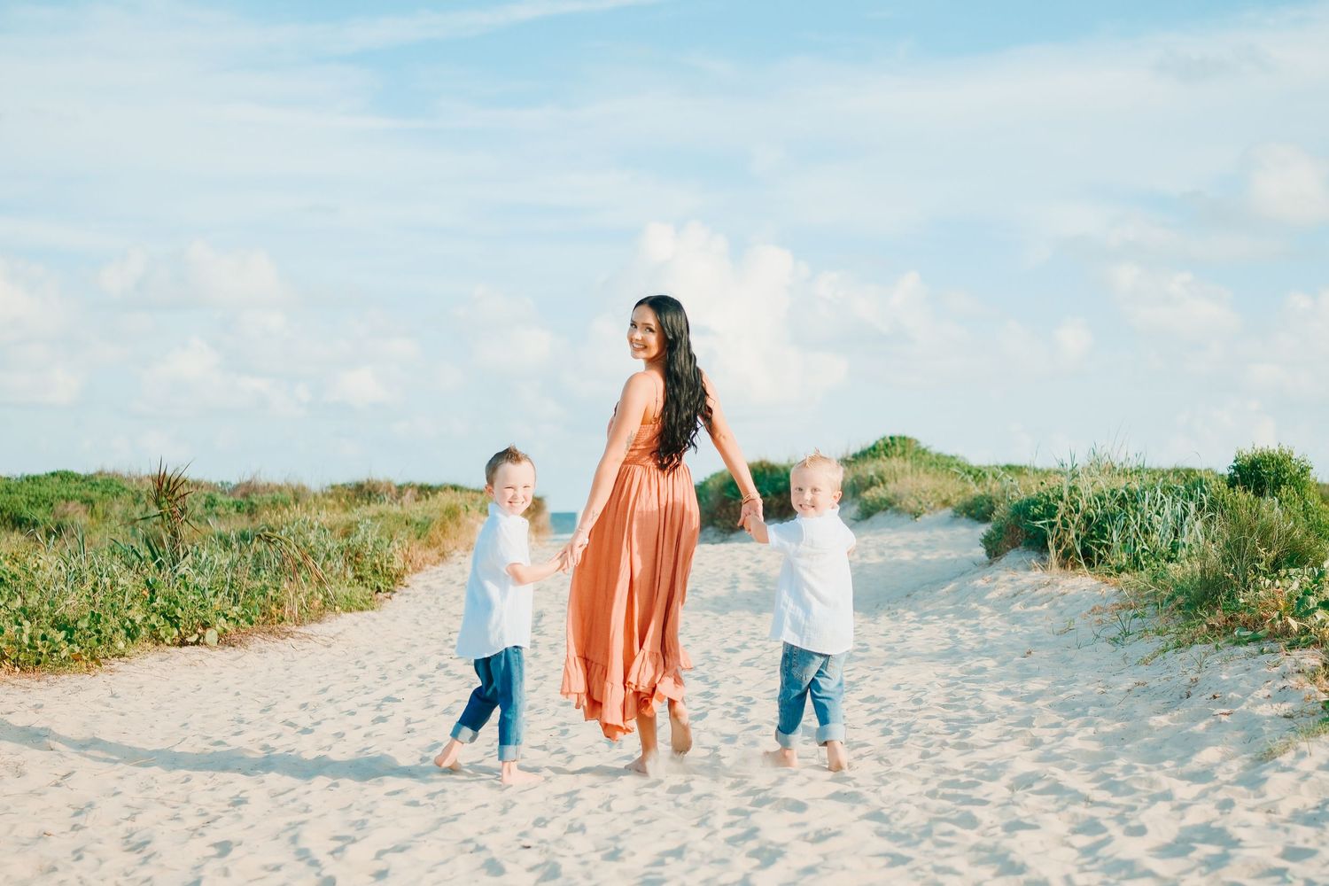A mother in an orange dress holds hands with two young children on a sandy beach path surrounded by dunes.