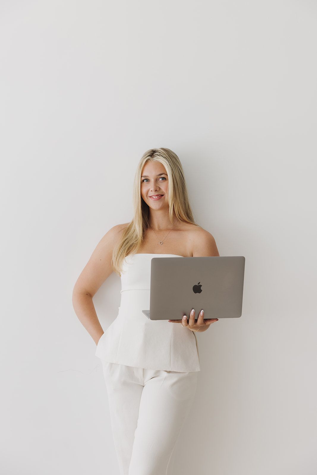 Smiling blonde woman in white strapless outfit holding an Apple MacBook laptop.