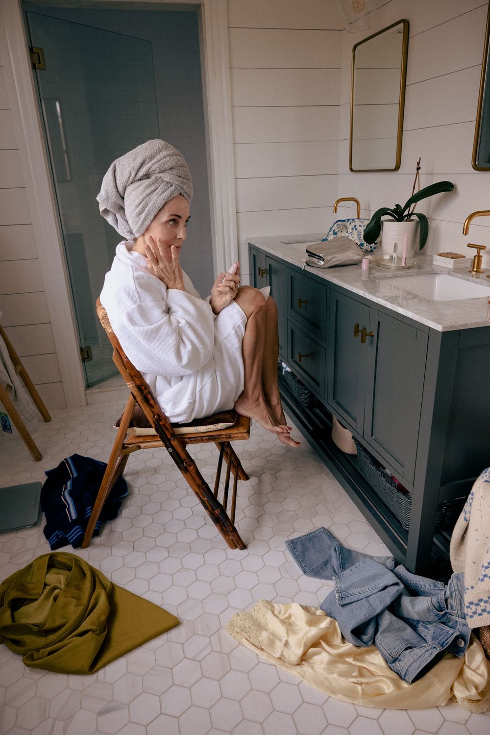 Woman in a white bathrobe and towel turban sitting in a bathroom, applying skincare near a dark green vanity.
