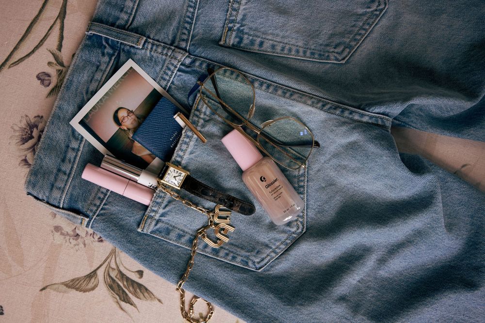 Flat lay of denim jeans with pink makeup products, sunglasses, a gold watch, and a Polaroid photo on fabric.