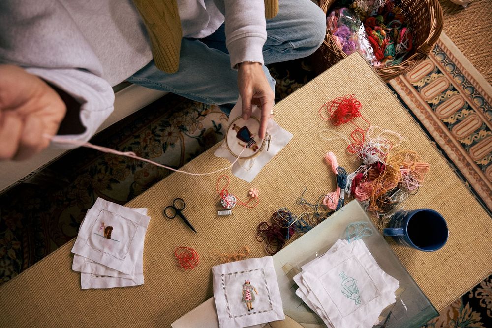 Overhead view of hands doing embroidery on white fabric, surrounded by colorful threads and craft supplies on a mat.